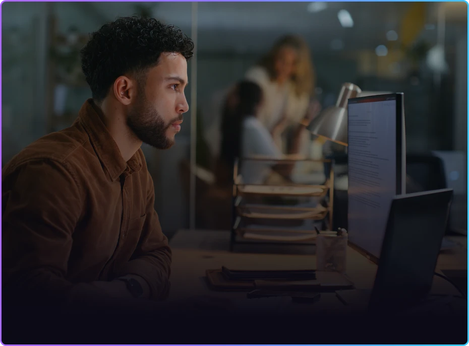 Man working late at a computer in an office with colleagues blurred in the background.